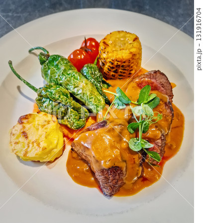A vibrant dish of sliced beef steak topped with rich orange sauce and garnished with fresh microgreens, fire-roasted corn, grilled padron peppers, charred cherry tomatoes and cheesy potato gratin 131916704