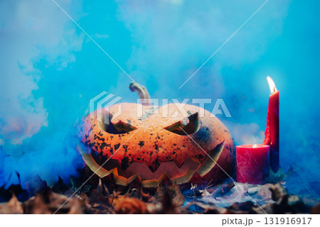 Pumpkin with carved grimace and candles next to it. Jack-o-lantern on orange foliage in blue smoke Pumpkin with carved grimace and candles next to it. Jack-o-lantern on orange foliage in blue smoke 131916917