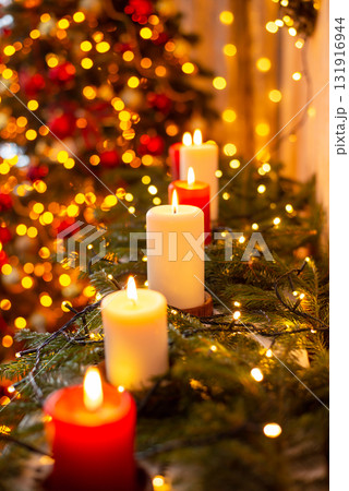 Vertical shot of line of burning candles in decorated room, Christmas tree on the background. Festive warm atmosphere at home Vertical shot of line of burning candles in decorated room, Christmas tree on the background. Festive warm atmosphere at home 131916944