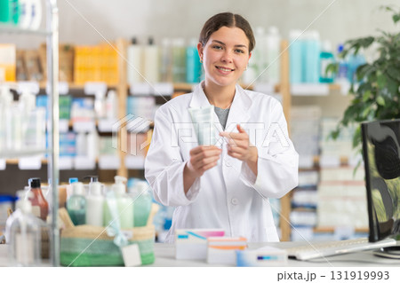 Polite experienced young girl pharmacist standing at counter and offering cream or skin care product in pharmacy 131919993