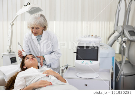 Young female visitor receiving hardware electric lifting massage with help of old female beautician in spa salon indoors Young female visitor receiving hardware electric lifting massage with help of old female beautician in spa salon indoors 131920285