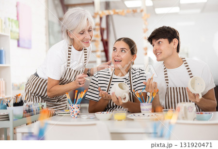 Senior woman assisting happy guy and girl painting ceramic mugs in workshop 131920471