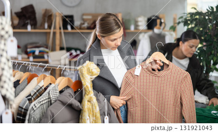 Positive young woman standing in interior of clothing boutique and choosing dress for autumn-winter season 131921043