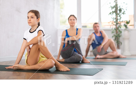 Middle-aged woman, man, teenage girl and boy practicing Lord of the Fishes Pose, or Matsyendrasana on mats during family Hatha yoga class in wellness studio 131921289