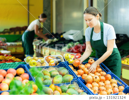 Positive saleswoman laying out pomegranates on display stand in supermarket Positive saleswoman laying out pomegranates on display stand in supermarket 131921324