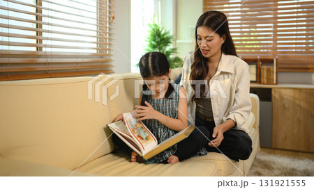 An Asian mother and daughter sit together indoors, smiling and bonding over a book An Asian mother and daughter sit together indoors, smiling and bonding over a book 131921555