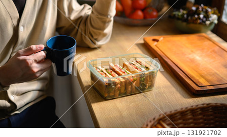 Morning breakfast scene with sandwiches in a lunchbox and a person holding a blue coffee mug on a kitchen counter 131921702