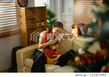 Happy mother sitting beside daughter using digital tablet in cozy living room with Christmas decorations 131921945