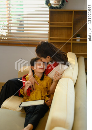 Loving mother kissing daughter forehead while relaxing together on sofa during Christmas season Loving mother kissing daughter forehead while relaxing together on sofa during Christmas season 131921948