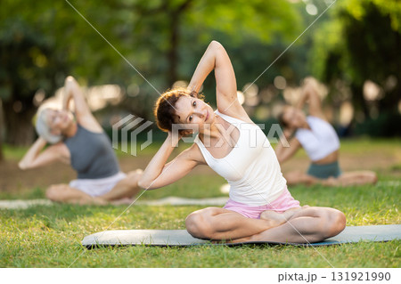 Class of calm sports women in activewear stretching muscles and doing tilts while sitting in lotus pose on mats during yoga training together at city park Class of calm sports women in activewear stretching muscles and doing tilts while sitting in lotus pose on mats during yoga training together at city park 131921990