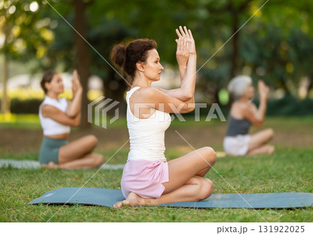 Calm european young female practicing Half Lord of fishes pose or Ardha Matsyendrasana during yoga training with group in green park Calm european young female practicing Half Lord of fishes pose or Ardha Matsyendrasana during yoga training with group in green park 131922025