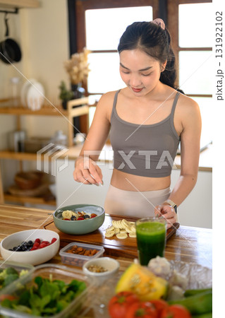 Woman in activewear preparing a healthy yogurt bowl topped with fresh fruits, nuts, and seeds for a nutritious breakfast. 131922102