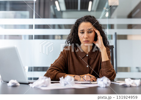 Stressed young woman dealing with a severe headache and emotional burnout, sitting alone at an office desk with crumpled paper and a laptop, struggling with performance pressure and job frustration 131924959