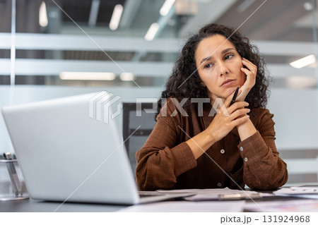 Young woman sitting at her office desk, looking tired and stressed, experiencing burnout from work while holding a pen and contemplating with hands on her face Young woman sitting at her office desk, looking tired and stressed, experiencing burnout from work while holding a pen and contemplating with hands on her face 131924968