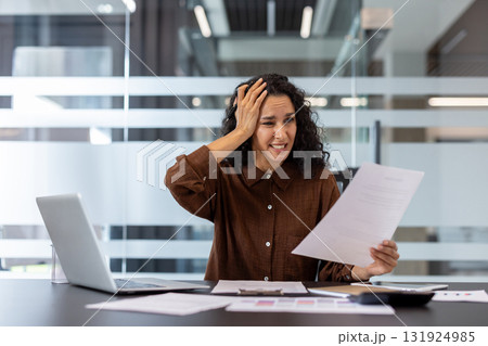 Stressed businesswoman holding a document and clutching her head, experiencing frustration and anxiety from work problems and overwhelming paperwork in her office cubicle 131924985