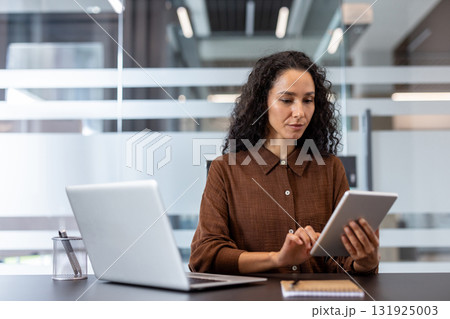 Professional businesswoman working at her desk, concentrating while using a digital tablet for tasks and checking information, with a laptop nearby in a contemporary office setting 131925003