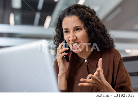 Woman expressing frustration and anger while talking loudly on a mobile phone in an office, her tense face and gesturing hand showing extreme stress and upset during a difficult argument 131925015