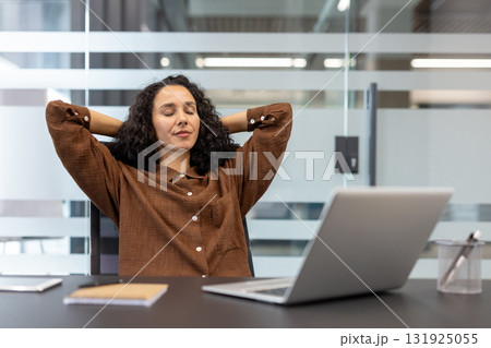 Young woman with curly hair leaning back at her desk, eyes closed and hands behind head, enjoying a calm, stress-free break during a busy remote workday at home 131925055