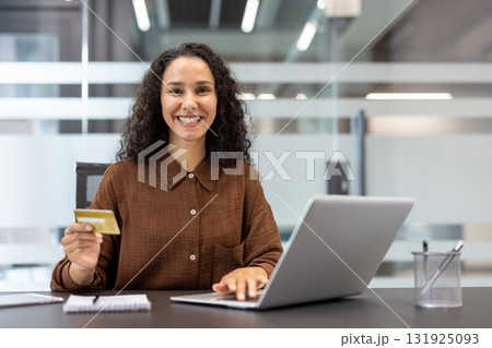 Woman smiling and holding a gold credit card, sitting at a desk and using a laptop for online shopping or e-commerce purchases in a modern office setup 131925093
