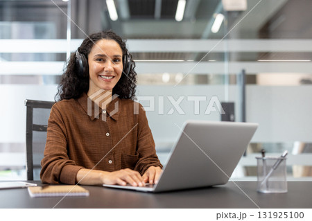 Professional woman wearing headphones smiling at the camera while typing on a laptop, engaged in remote work or online meeting from a modern office environment Professional woman wearing headphones smiling at the camera while typing on a laptop, engaged in remote work or online meeting from a modern office environment 131925100