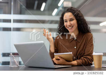 Young businesswoman smiling during a remote video call on a laptop, explaining a concept and taking notes, while working from a modern corporate office Young businesswoman smiling during a remote video call on a laptop, explaining a concept and taking notes, while working from a modern corporate office 131925102