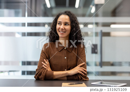 Confident young executive woman with curly hair sitting at an office desk, smiling and looking away, embodying success, professionalism, and positive thinking in a modern workplace environment 131925109