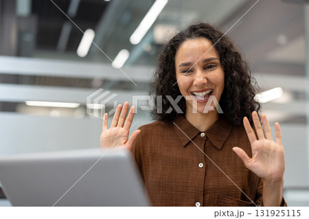 Woman engaging in a video call and happily waving her hands, communicating and participating in an online meeting with a laptop from a modern office environment 131925115