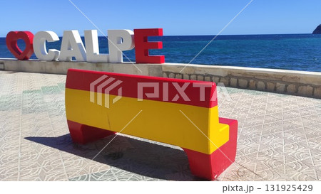 Colorful bench painted in red and yellow sits on a tiled promenade overlooking the blue sea, with large letters spelling a location in the background, creating a vibrant coastal scene 131925429