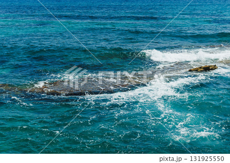 Waves crash against rocky shoreline under bright blue sky, capturing the tranquil beauty of the ocean at midday 131925550
