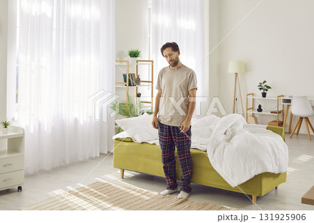 Young sleepy man suffering from sleepwalking standing with closed eyes near his bed at home. 131925906
