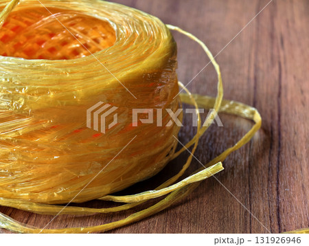 Close-up of yellow raffia plastic rope roll on wooden surface. Strong, durable, and flexible cord used for packaging, shipping, agriculture, DIY, and craft projects. 131926946