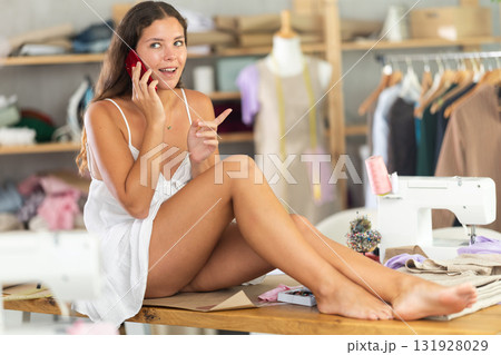 Barefoot female dressmaker sitting on table chatting on phone Barefoot female dressmaker sitting on table chatting on phone 131928029