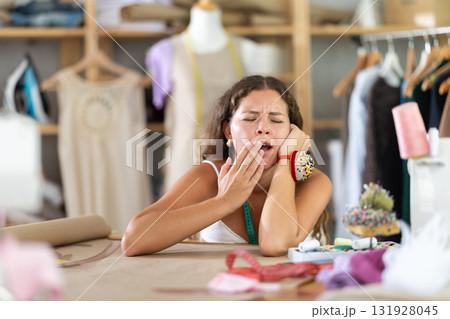 Exhausted young seamstress yawning at worktable 131928045