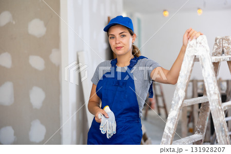 Portrait of positive builder woman in blue overalls next to stepladder 131928207