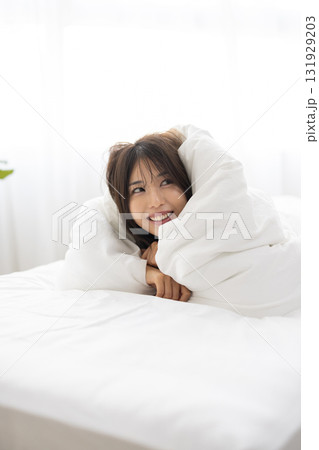 朝日を浴びながら布団をかぶって微笑む若い女性 131929203