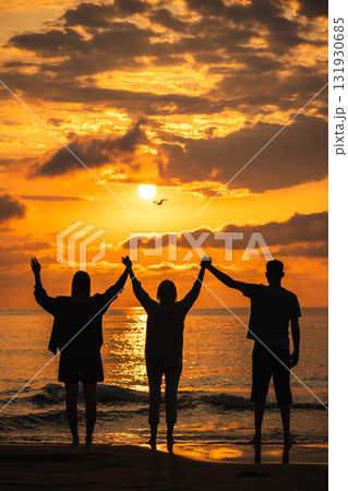 Three people standing on the beach at sunrise with raised arms, watching a flying seagull above the glowing horizon 131930685