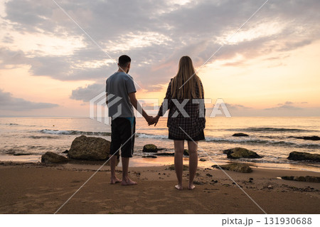 Couple holding hands and looking at the sunrise over the calm sea, standing barefoot on a sandy beach with rocks and soft morning light reflecting on the water 131930688