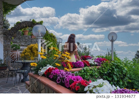 A terrace in Amalfi, Italy, features vibrant flowers, a stone archway, wrought iron furniture, and a woman in a yellow dress under a cloudy sky. A terrace in Amalfi, Italy, features vibrant flowers, a stone archway, wrought iron furniture, and a woman in a yellow dress under a cloudy sky. 131934677