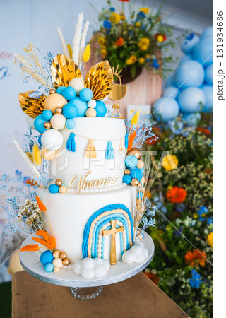 A white three tiered cake with blue, gold, and orange accents, featuring a gold cross, floral arrangements, blue balloons, and the name Vincenzo. A white three tiered cake with blue, gold, and orange accents, featuring a gold cross, floral arrangements, blue balloons, and the name Vincenzo. 131934686