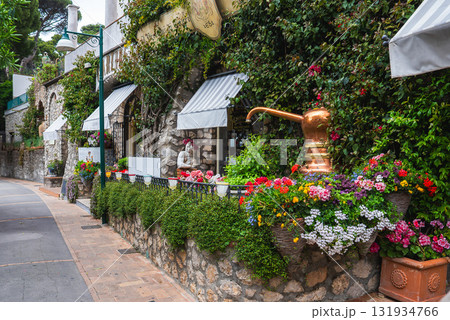 A stone building on Capri, Italy, features lush greenery, vibrant flowers, striped awnings, a brass spout, cobblestone sidewalk, and rustic walls. A stone building on Capri, Italy, features lush greenery, vibrant flowers, striped awnings, a brass spout, cobblestone sidewalk, and rustic walls. 131934766