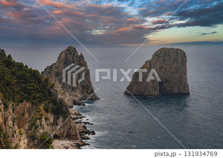 The Faraglioni rock formations rise from the Tyrrhenian Sea near Capri, Italy, with rugged cliffs and a vibrant sunset sky of orange, pink, and blue. 131934769