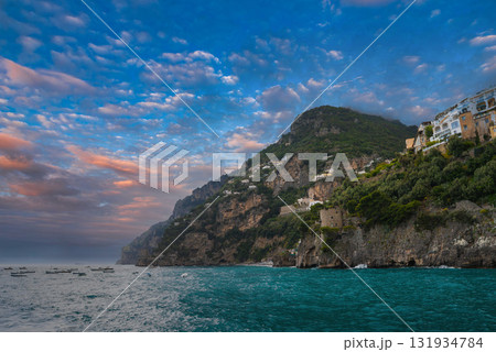 Positano, Italy, with pastel buildings on steep cliffs, lush greenery, and boats on turquoise waters under a colorful sunset sky. 131934784