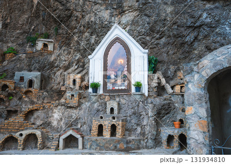 A carved rocky cliff features an arched frame with painted artwork, flanked by potted plants and miniature stone structures in Positano, Italy. 131934810