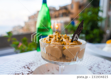 A creamy dessert in a glass dish topped with pistachios and chocolate, set against blurred terraced buildings and greenery in Positano, Italy. A creamy dessert in a glass dish topped with pistachios and chocolate, set against blurred terraced buildings and greenery in Positano, Italy. 131934815