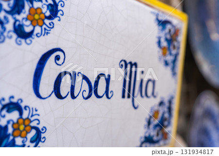 A ceramic tile with Casa Mia in blue script, featuring floral patterns in blue and yellow. The cracked glaze highlights Italian craftsmanship in Ravello. 131934817