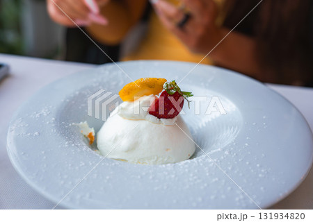 A dome of whipped cream or mousse topped with a strawberry and orange slice on a white plate, dusted with powdered sugar in a casual dining setting. A dome of whipped cream or mousse topped with a strawberry and orange slice on a white plate, dusted with powdered sugar in a casual dining setting. 131934820