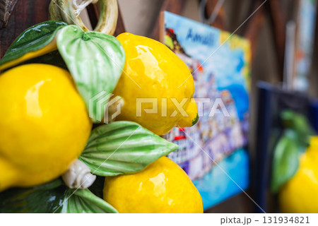 Vibrant ceramic lemons with green leaves are displayed in the foreground, with a colorful painting of a coastal town, likely Amalfi, in the background. 131934821