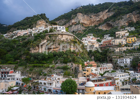 View of Positano, Italy, with vibrant buildings on a steep hillside, a rocky outcrop with vegetation, and an overcast sky creating a dramatic scene. 131934824