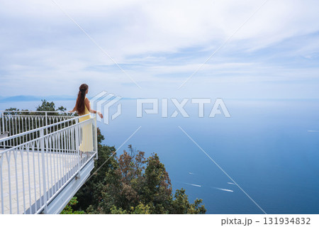 A beautiful woman in a yellow dress stands on a white balcony above lush greenery, overlooking the Tyrrhenian Sea with boats and distant landforms in view. 131934832