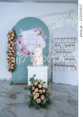 Elegant first communion setup with a pastel green backdrop, floral cake on a white pedestal, peach roses, party favors, and ornate white wall detailing. Elegant first communion setup with a pastel green backdrop, floral cake on a white pedestal, peach roses, party favors, and ornate white wall detailing. 131934837